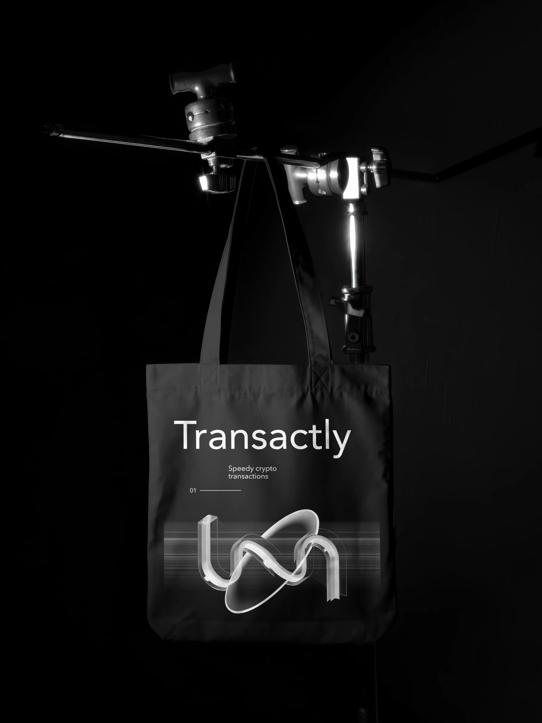 A black tote bag on a light stand, printed with the Transactly wordmark and the glass mark over speed lines